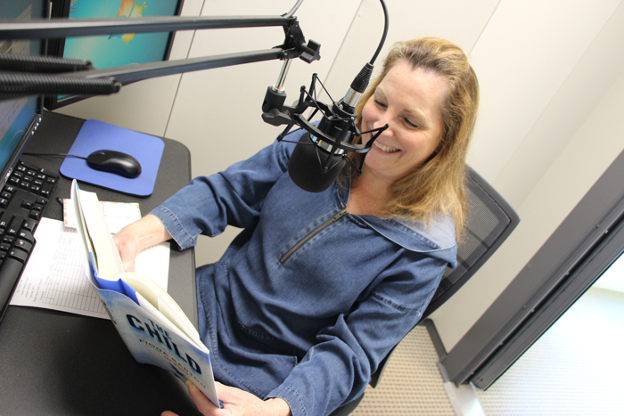 Woman wearing a blue sweatshirt sits in a recording booth reading The Child into a microphone.