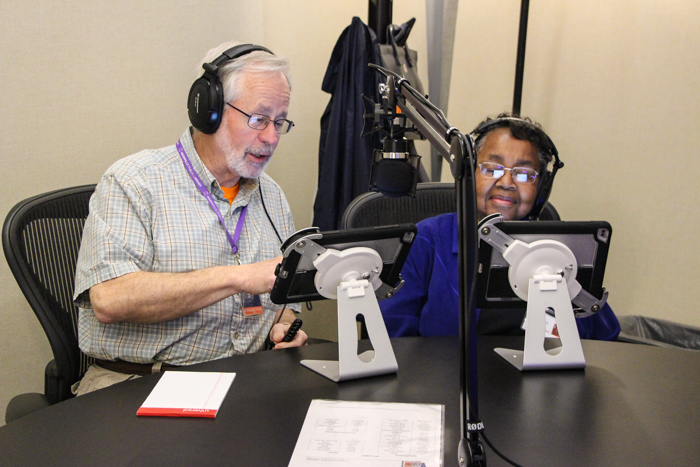 A man and a woman sit in a recording booth reading into a microphone.